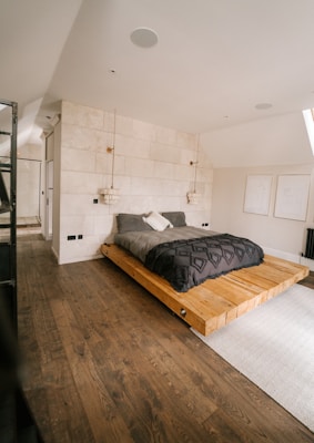 A modern bedroom with a minimalist design featuring a low wooden platform bed. The bedding consists of dark gray and white pillows, set against a textured white stone wall. Two pendant lights hang from the ceiling, adding a touch of elegance. The room includes light wooden flooring and simple line art on the walls. Natural light enters from a window, creating a warm, serene atmosphere.