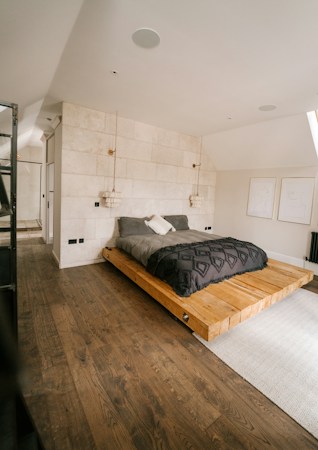 A modern bedroom with a minimalist design featuring a low wooden platform bed. The bedding consists of dark gray and white pillows, set against a textured white stone wall. Two pendant lights hang from the ceiling, adding a touch of elegance. The room includes light wooden flooring and simple line art on the walls. Natural light enters from a window, creating a warm, serene atmosphere.