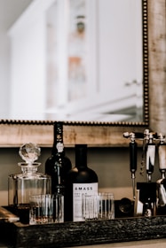 A tabletop display featuring a collection of barware items, including a liquor bottle labeled 'Amass,' a crystal decanter filled with a dark liquid, and several glass tumblers. A bottle of port and a set of cocktail-making tools, such as a corkscrew and muddler, are also present. The scene is reflected in a large mirror with a textured wooden frame that captures part of a blurred background, suggesting a home or kitchen setting.