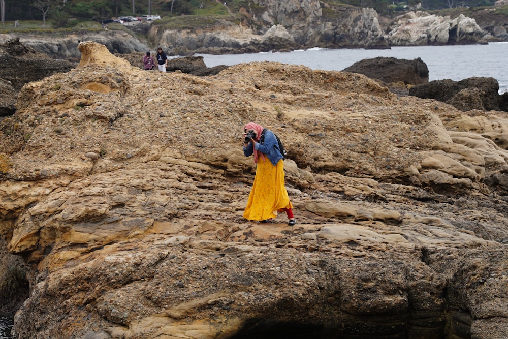 woman in yellow dress standing on brown rock near body of water during daytime