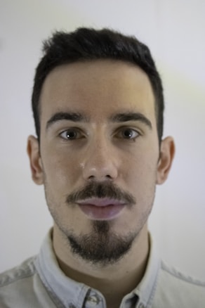 A portrait of a person with short dark hair, facial hair, and a neutral expression, wearing a light-colored collared shirt. The background is softly blurred.