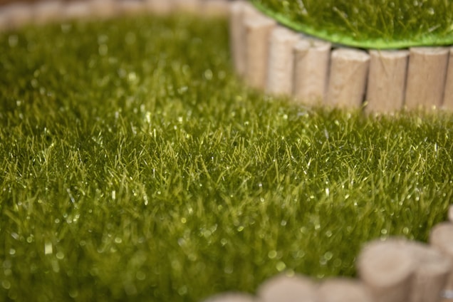 A close-up view of a patch of artificial grass bordered by small wooden posts. The texture of the synthetic grass is evident, showing fine, green blades with a slight sheen under the light.