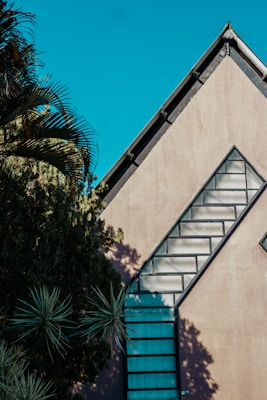 The image features a modern architectural structure with a distinct triangular roofline. A series of rectangular windows are arranged diagonally along the side of the building, creating an artistic pattern. Lush greenery, including palm leaves, adds a natural element and casts intriguing shadows on the building's surface. The sky is clear, providing a vibrant backdrop.