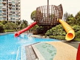 A modern outdoor play area featuring a unique, spherical wooden structure with two twisty slides, one red and one yellow, extending into a clear blue swimming pool. Surrounding the play structure are lush green plants and well-maintained pathways. In the background, a multi-story building with balconies can be seen, indicating an urban setting.