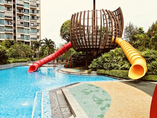 A modern outdoor play area featuring a unique, spherical wooden structure with two twisty slides, one red and one yellow, extending into a clear blue swimming pool. Surrounding the play structure are lush green plants and well-maintained pathways. In the background, a multi-story building with balconies can be seen, indicating an urban setting.