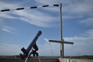 A telescope mounted on a tripod is positioned on a wooden deck, pointing towards the sky. A windsock and a wooden cross stand in the background against a blue sky with scattered white clouds. The horizon shows a grassy landscape and the sea.