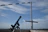 A telescope mounted on a tripod is positioned on a wooden deck, pointing towards the sky. A windsock and a wooden cross stand in the background against a blue sky with scattered white clouds. The horizon shows a grassy landscape and the sea.