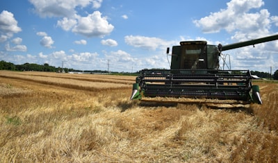 A large, green harvester is working in a vast field of golden wheat under a partly cloudy blue sky. The harvester is positioned towards the center-right of the image, capturing and processing the crops. Rolling fields and a row of trees can be seen in the background, along with several power lines stretching across the horizon.