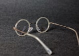 Round vintage-style eyeglasses with thin metal frames resting on a dark textured surface. The lenses are clear, and the glasses are positioned at an angle.