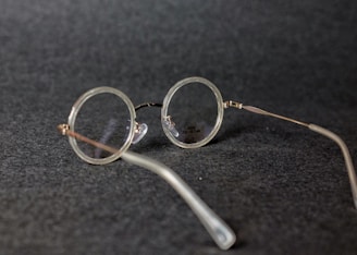 Round vintage-style eyeglasses with thin metal frames resting on a dark textured surface. The lenses are clear, and the glasses are positioned at an angle.