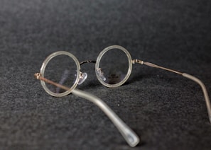 Round vintage-style eyeglasses with thin metal frames resting on a dark textured surface. The lenses are clear, and the glasses are positioned at an angle.