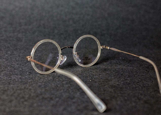 Round vintage-style eyeglasses with thin metal frames resting on a dark textured surface. The lenses are clear, and the glasses are positioned at an angle.