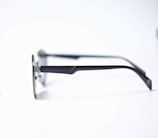 A pair of eyeglasses with sleek, dark metal frames and thin temples resting on a white surface. The glasses have a minimalist design with a slight curve at the temple tips.