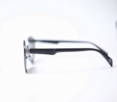 A pair of eyeglasses with sleek, dark metal frames and thin temples resting on a white surface. The glasses have a minimalist design with a slight curve at the temple tips.