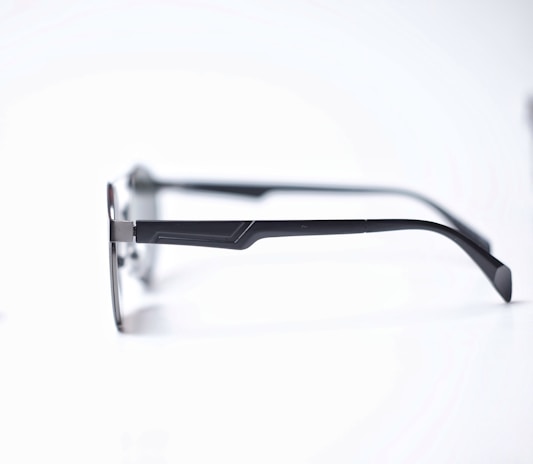 A pair of eyeglasses with sleek, dark metal frames and thin temples resting on a white surface. The glasses have a minimalist design with a slight curve at the temple tips.