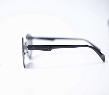 A pair of eyeglasses with sleek, dark metal frames and thin temples resting on a white surface. The glasses have a minimalist design with a slight curve at the temple tips.