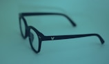 A pair of black eyeglasses with square frames, placed against a smooth, monochromatic blue-green background. The glasses have a small, white logo on the arm.