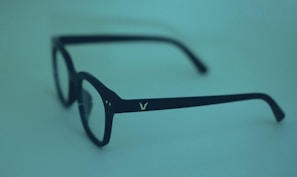 A pair of black eyeglasses with square frames, placed against a smooth, monochromatic blue-green background. The glasses have a small, white logo on the arm.
