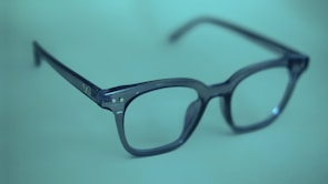 A pair of dark-rimmed eyeglasses rests on a soft, blurred green background. The glasses have a simple, classic design with a rectangular frame and clear lenses. The lighting casts a subtle shadow, adding depth to the image.