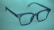 A pair of dark-rimmed eyeglasses rests on a soft, blurred green background. The glasses have a simple, classic design with a rectangular frame and clear lenses. The lighting casts a subtle shadow, adding depth to the image.