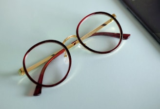 A pair of round eyeglasses with brown frames and gold temples is lying on a light blue surface. The glasses' lenses are clear, and a portion of a black object, possibly a mobile phone or case, is visible on the right edge of the image.