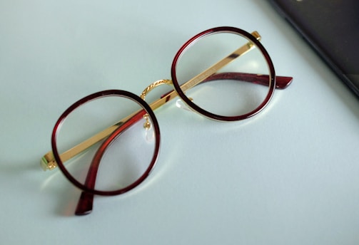 A pair of round eyeglasses with brown frames and gold temples is lying on a light blue surface. The glasses' lenses are clear, and a portion of a black object, possibly a mobile phone or case, is visible on the right edge of the image.