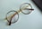 A pair of round eyeglasses with brown frames and gold temples is lying on a light blue surface. The glasses' lenses are clear, and a portion of a black object, possibly a mobile phone or case, is visible on the right edge of the image.