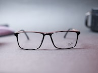 A pair of Ray-Ban eyeglasses with a sleek, rectangular frame is centered on a soft-focus surface. The muted background features hints of blurred shapes and colors, conveying a minimalist and subtle aesthetic.
