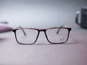 A pair of Ray-Ban eyeglasses with a sleek, rectangular frame is centered on a soft-focus surface. The muted background features hints of blurred shapes and colors, conveying a minimalist and subtle aesthetic.