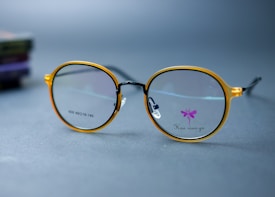 Round eyeglasses with a thin, golden frame are placed on a smooth, gray surface. The glasses have clear lenses, one of which shows a small purple butterfly design and text printed near the bottom. The background includes a blurred stack of colored books, adding depth to the image.