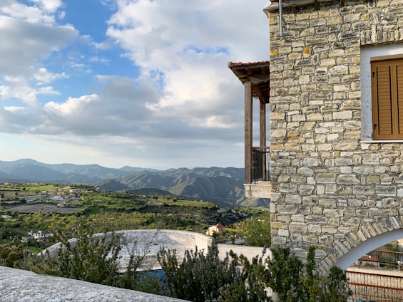 A stone house with a balcony and wooden shutters sits prominently in the foreground on the right. In the background, there is a vast landscape of rolling green hills and mountains under a partly cloudy sky. The area has scattered houses, vegetation, and appears tranquil and scenic.