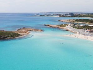 A picturesque coastal scene featuring clear turquoise waters and a sandy beach. The beach is populated with sun umbrellas and lounging chairs, while a few people can be seen enjoying the sun and swimming. In the distance, a hotel and other buildings are visible, along with lush greenery and palm trees lining the shore.
