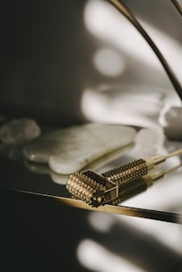 A textured, gold-toned massage tool with small spikes is placed on a reflective surface. In the background, there are smooth, white stones or crystals. The lighting creates soft, diffused shadows that add a serene and luxurious feel.