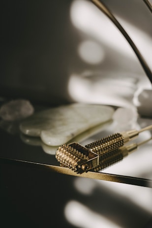 A textured, gold-toned massage tool with small spikes is placed on a reflective surface. In the background, there are smooth, white stones or crystals. The lighting creates soft, diffused shadows that add a serene and luxurious feel.