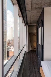 A modern interior corridor of a building features a large window wall extending to the floor, offering a view of a cityscape with tall buildings and blue sky. The corridor has a dark wooden floor, a bare concrete ceiling, and a sliding door leading to a space with minimalistic wooden furniture, including a hanging rack and a chair.