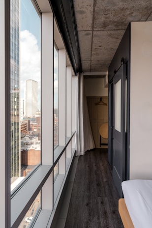 A modern interior corridor of a building features a large window wall extending to the floor, offering a view of a cityscape with tall buildings and blue sky. The corridor has a dark wooden floor, a bare concrete ceiling, and a sliding door leading to a space with minimalistic wooden furniture, including a hanging rack and a chair.
