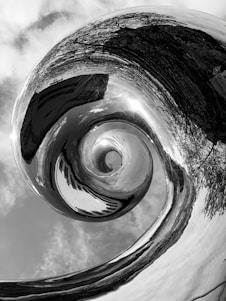 A black and white abstract image featuring a swirling, spiral vortex. The vortex appears to distort its surroundings, including reflections of trees and sky. The spiral creates a dynamic visual effect, drawing the viewer's eye towards the center.