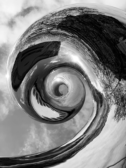 A black and white abstract image featuring a swirling, spiral vortex. The vortex appears to distort its surroundings, including reflections of trees and sky. The spiral creates a dynamic visual effect, drawing the viewer's eye towards the center.
