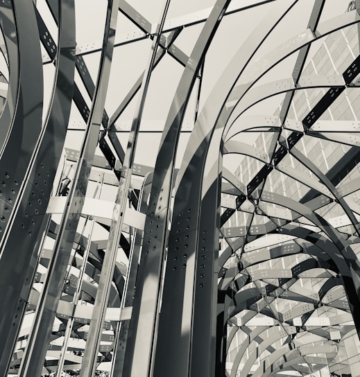 An intricate network of arching metal beams, intersecting at various angles, creates a complex and dynamic architectural pattern. The structure appears to be part of a modern building facade, with a grid-like windowed wall visible in the background. The light casts geometric shadows, enhancing the spatial depth.