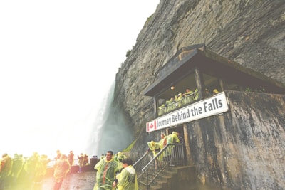 People wearing yellow ponchos stand near a rocky cliff with a building that has a sign reading 'Journey Behind the Falls'. Water cascades down a massive waterfall beside them.