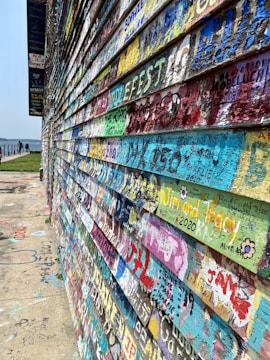 A wall covered with colorful graffiti and messages in various fonts and sizes. The surface consists of wooden panels with inscriptions in vibrant shades of blue, red, yellow, and green. The background shows an outdoor setting with a walkway leading to a distant railing and a view of water.