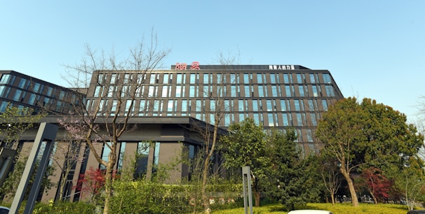 A modern office building features a sleek design with large glass windows and clean lines. Surrounding the building are various trees and shrubs, creating a green and natural environment. The sky is clear and blue, adding to the overall tranquil setting.