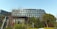 A modern office building features a sleek design with large glass windows and clean lines. Surrounding the building are various trees and shrubs, creating a green and natural environment. The sky is clear and blue, adding to the overall tranquil setting.