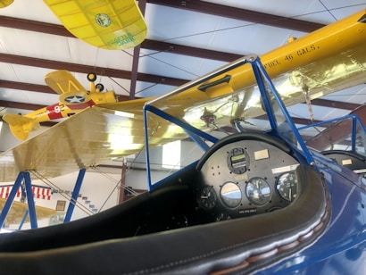 The image displays a cockpit interior of an old aircraft with detailed gauges and a leather-trimmed dashboard. Overhead, two vintage biplanes with yellow wings are suspended in a hangar. The planes feature military markings and insignia, and the wing of one shows a 'FUEL 46 GALS.' label.