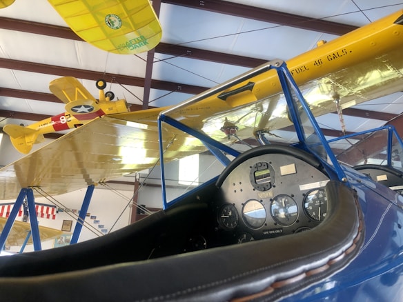 The image displays a cockpit interior of an old aircraft with detailed gauges and a leather-trimmed dashboard. Overhead, two vintage biplanes with yellow wings are suspended in a hangar. The planes feature military markings and insignia, and the wing of one shows a 'FUEL 46 GALS.' label.