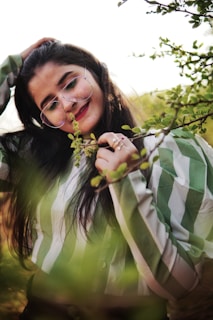 A woman with long dark hair is wearing round glasses and a striped green and white blouse, standing amidst lush green foliage. She is smiling gently, with one hand touching her hair and the other holding a sprig of leaves.
