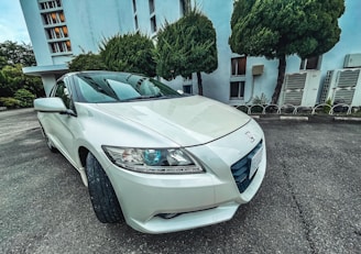 A white Honda car is parked in front of a modern building with large windows. The vehicle is positioned at an angle, highlighting its front and side features. Surrounding the scene are well-maintained trees and shrubs.