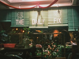A cozy cafe setting with a sign displaying 'The Daily Fix' in bold letters. The interior features a warm, inviting atmosphere with green bamboo blinds and soft lighting. The foreground includes blurred plants and decorative elements, enhancing the intimate feel of the space.