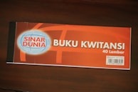 A receipt book labeled 'Buku Kwitansi' with 40 sheets, featuring the brand 'Sinar Dunia'. The cover is predominantly orange with a blue and white globe logo on the left. A barcode is visible on the bottom right corner, along with some additional information.