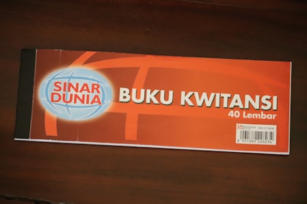 A receipt book labeled 'Buku Kwitansi' with 40 sheets, featuring the brand 'Sinar Dunia'. The cover is predominantly orange with a blue and white globe logo on the left. A barcode is visible on the bottom right corner, along with some additional information.